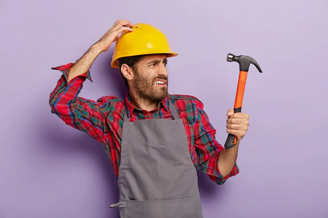 Foto eines unzufriedenen Handwerkers, der sich nach rechts dreht, mit gerunzelter Stirn in die Ferne blickt, einen Hammer hält, als professioneller Bauarbeiter tätig ist, ein neues zu reparierendes Objekt bemerkt und Helm sowie Schürze trägt. Renovierung, Ingenieurwesen.